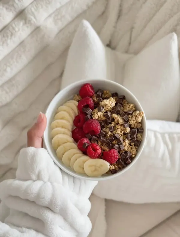 Tigela de granola com banana e framboesas nas mãos de uma mulher de roupão