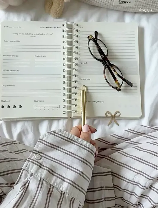 Caderno de bem-estar aberto sobre a cama, com óculos, caneta dourada e pelúcia